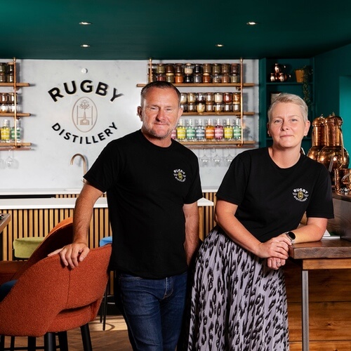 a man and woman standing in front of the rugby distillery sign in a club house